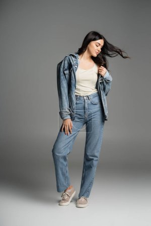 Photo for Full length of charming woman with long brunette hair waving from wind posing in stylish blue jeans and denim jacket while standing on grey background - Royalty Free Image