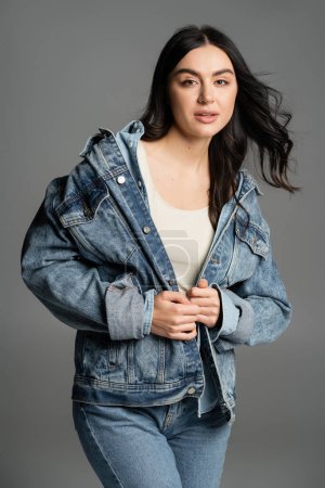 charming young woman with brunette long hair posing in fashionable and blue denim jacket looking at camera while standing on grey background