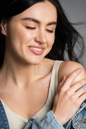 Photo for Portrait of alluring woman with brunette hair posing in stylish and blue denim jacket and holding hand near shoulder while smiling with closed eyes isolated on grey background - Royalty Free Image
