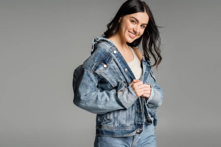 portrait of cheerful young woman with brunette hair posing in stylish denim jacket and blue jeans while smiling and standing isolated on grey background