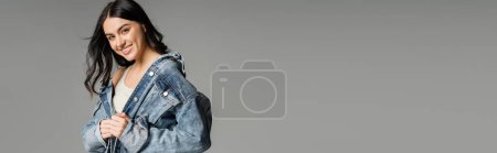 Photo for Portrait of cheerful young woman with brunette and gorgeous hair posing in stylish denim jacket while smiling and standing isolated on grey background, banner - Royalty Free Image