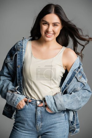 happy young woman with brunette hair standing with hands in pockets of blue jeans and posing in stylish denim jacket while smiling isolated on grey background