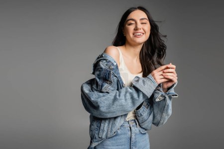 Photo for Positive young woman with brunette hair standing in blue jeans and fashionable denim jacket while smiling and posing isolated on grey background - Royalty Free Image