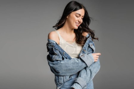 Photo for Alluring young woman with brunette hair and natural makeup standing in blue jeans and fashionable denim jacket while smiling and posing isolated on grey background - Royalty Free Image