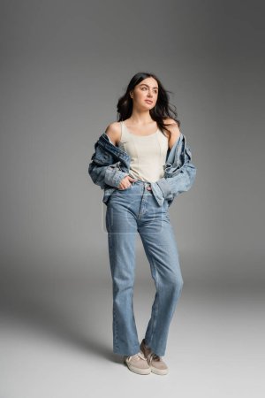 full length of young charming woman with long brunette hair posing in stylish blue jeans and denim jacket while standing with hands in pockets on grey background