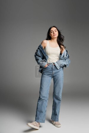 full length of young appealing woman with long brunette hair posing in stylish blue jeans and denim jacket while standing with hands in pockets on grey background