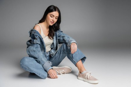 full length of young charming woman with long brunette hair and flawless natural makeup posing in stylish blue jeans and denim jacket while sitting on grey background