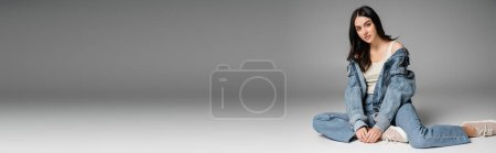 Photo for Full length of enchanting model with long brunette hair and flawless natural makeup posing in stylish blue jeans and denim jacket while sitting on grey background, banner - Royalty Free Image