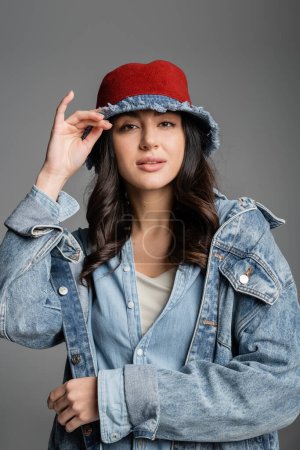 portrait of young alluring woman with flawless natural makeup posing in panama hat and denim jacket and looking at camera on grey background