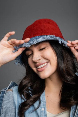 retrato de mujer feliz con los ojos cerrados y maquillaje natural impecable posando en sombrero de panama y chaqueta de mezclilla y sonriendo sobre fondo gris
