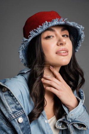 portrait of young charming woman with flawless natural makeup posing in panama hat and denim jacket and looking at camera on grey background