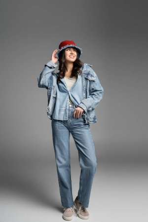 full length of young happy woman with flawless natural makeup adjusting denim panama hat and posing in blue jeans and jacket while standing and looking away on grey background