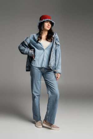 full length of young charming woman with flawless natural makeup posing in denim panama hat, blue jeans and jacket while standing with hand on hip on grey background
