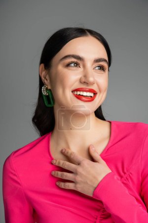 portrait of happy young woman with shiny brunette hair, trendy earrings, red lips and stylish magenta dress smiling while looking away and posing isolated on grey background 