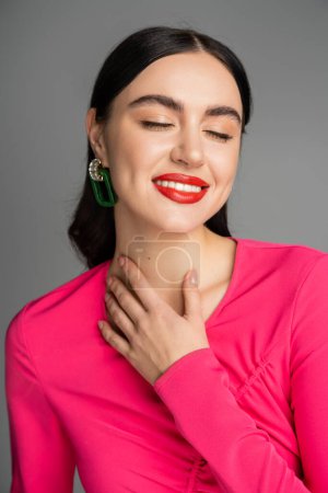 portrait of pleased woman with shiny brunette hair, trendy earrings, red lips and stylish magenta dress smiling with closed eyes isolated on grey background 