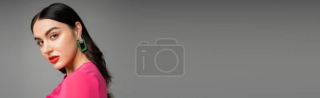 Charmante junge Frau mit brünetten Haaren, trendigen Ohrringen, roten Lippen und stylischem Magentakleid, die in die Kamera blickt und auf grauem Hintergrund posiert, Banner 