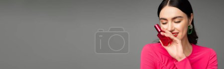 Téléchargez les photos : Femme glamour avec cheveux bruns, boucles d'oreilles tendance, lèvres rouges et élégante robe magenta tenant bouteille de parfum luxueux et souriant sur fond gris, bannière - en image libre de droit