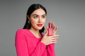 alluring young woman with brunette hair, trendy earrings, red lips and stylish magenta dress holding bottle of luxurious perfume isolated on grey background  Longsleeve T-shirt #654499520