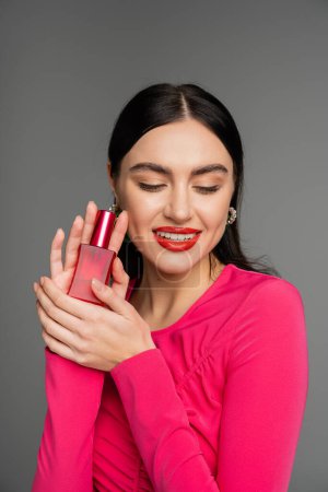Porträt einer wunderschönen jungen Frau mit brünetten Haaren und roten Lippen, die in einem magentafarbenen Kleid posiert, während sie eine Flasche luxuriösen Parfüms in der Hand hält und isoliert auf grauem Hintergrund lächelt 