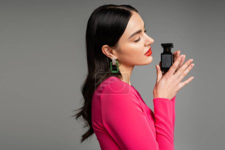 Photo for Side view of alluring woman with shiny brunette hair, red lips and closed eyes posing in trendy magenta dress while holding bottle of luxurious perfume isolated on grey background - Royalty Free Image