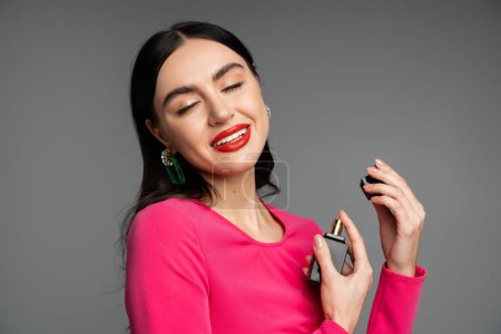 sophisticated young woman with brunette hair, red lips, closed eyes and magenta dress holding bottle and spraying luxurious perfume while smiling on grey background  magic mug #654499684