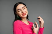 sophisticated young woman with brunette hair, red lips, closed eyes and magenta dress holding bottle and spraying luxurious perfume while smiling on grey background  Stickers #654499684