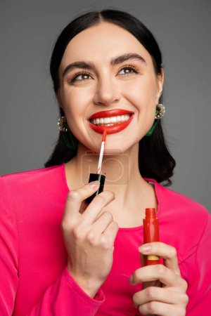 alluring young woman with shiny brunette hair and trendy earrings posing in magenta dress while applying red lip gloss on lips and smiling isolated on grey background 