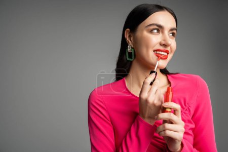 Charmante junge Frau mit brünetten Haaren und trendigen Ohrringen posiert im magentafarbenen Kleid, während sie roten Lipgloss auf die Lippen aufträgt und isoliert auf grauem Hintergrund lächelt 