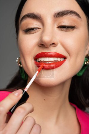 close up view of enchanting young woman with brunette hair and trendy earrings applying red lip gloss and smiling before party isolated on grey background 