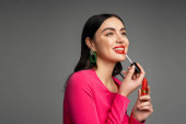 happy woman with brunette hair and trendy earrings applying red lip gloss and smiling while posing in magenta dress and preparing for party isolated on grey background  mug #654499876