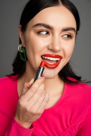 charming woman with trendy earrings and shiny brunette hair applying red lipstick and smiling while looking away and and preparing for party isolated on grey background mug #654499938