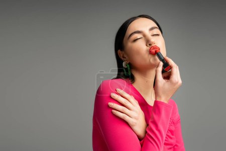 Foto de Mujer divertida con pendientes de moda y pelo moreno brillante aplicando lápiz labial rojo y labios pucheros mientras se prepara para la fiesta aislada sobre fondo gris - Imagen libre de derechos