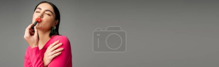 Foto de Mujer divertida con pendientes de moda y pelo moreno brillante aplicando lápiz labial rojo y labios pucheros mientras posa aislado sobre fondo gris, pancarta - Imagen libre de derechos