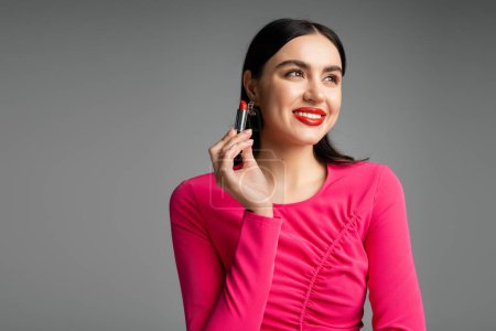 glückliche Frau mit trendigen Ohrringen und glänzenden brünetten Haaren, die roten Lippenstift hält und lächelt, während sie wegschaut und isoliert auf grauem Hintergrund posiert