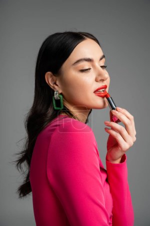 young and charming woman with trendy earrings and shiny brunette hair posing while applying red lipstick and looking away isolated on grey background