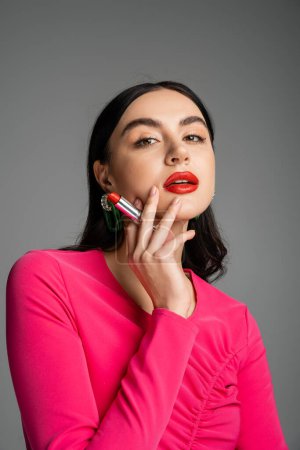 gorgeous woman with trendy earrings and shiny brunette hair holding red lipstick between fingers and looking at camera while posing isolated on grey background