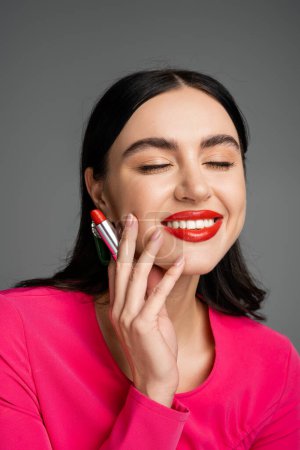 portrait of pretty woman with brunette hair holding red lipstick and smiling with closed eyes while posing in party dress isolated on grey background