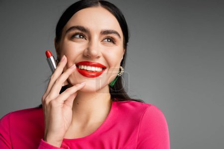 retrato de mujer encantadora con pendientes de moda y pelo moreno brillante sosteniendo lápiz labial rojo entre los dedos y sonriendo mientras posando sobre fondo gris 