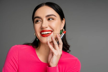 portrait of elegant woman with trendy earrings and shiny brunette hair holding red lipstick between fingers and smiling while posing on grey background 