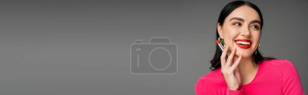 Foto de Retrato de mujer encantadora con pendientes de moda y cabello moreno sosteniendo lápiz labial rojo entre los dedos y sonriendo mientras posando sobre fondo gris, pancarta - Imagen libre de derechos