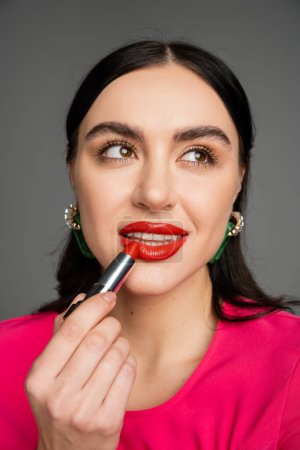 portrait of stunning young woman with trendy earrings and flawless makeup applying red lipstick while posing and looking away on grey background 