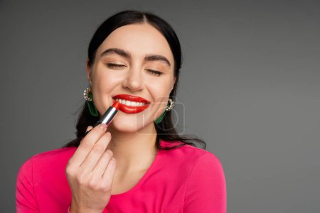 Foto de Retrato de mujer joven elegante con pendientes de moda y maquillaje impecable aplicando lápiz labial rojo y sonriendo con los ojos cerrados mientras posando sobre fondo gris - Imagen libre de derechos