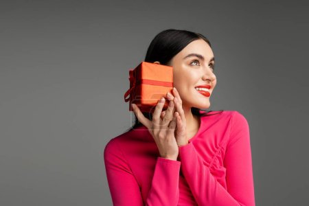 enchanting and chic young woman with trendy earrings and shiny brunette hair smiling while holding red and wrapped gift box on grey background with copy space 
