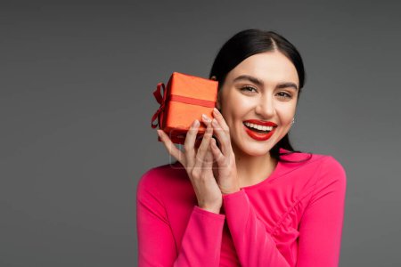 mujer joven positiva y elegante con pendientes de moda y cabello moreno brillante sonriendo mientras sostiene la caja de regalo roja y envuelta en fondo gris 