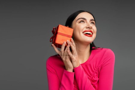 Photo for Happy young woman with trendy earrings and shiny brunette hair smiling while holding red and wrapped gift box on holiday and looking up on grey background - Royalty Free Image