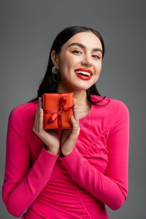 positive und junge Frau mit trendigen Ohrringen und brünetten Haaren, die lächelt, während sie rotes und eingewickeltes Geschenk für den Urlaub auf grauem Hintergrund hält 