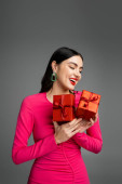 cheerful young woman with shiny brunette hair and trendy earrings smiling while standing in magenta party dress and holding wrapped gift boxes for holiday on grey background  hoodie #654500462