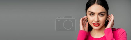 portrait de femme élégante et séduisante aux cheveux bruns, lèvres rouges et boucles d'oreilles tendance debout en robe de soirée magenta tout en regardant la caméra isolée sur fond gris, bannière 