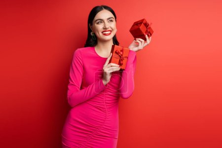 charming woman with brunette hair and trendy earrings smiling while standing in magenta party dress and holding wrapped gift boxes for holiday on red background 