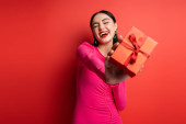 excited woman with brunette hair and trendy earrings smiling while standing in magenta party dress and holding wrapped gift box for holiday on red background  hoodie #654500818